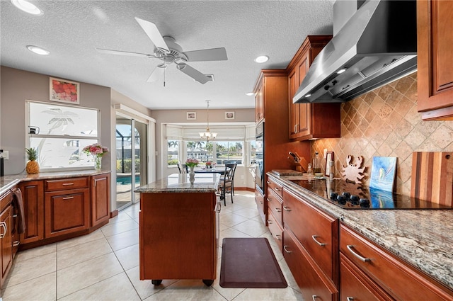 kitchen with light stone countertops, black appliances, a center island, decorative light fixtures, and wall chimney range hood