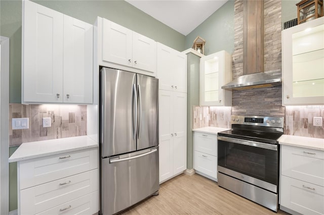 kitchen with wall chimney range hood, decorative backsplash, light wood-type flooring, appliances with stainless steel finishes, and white cabinetry