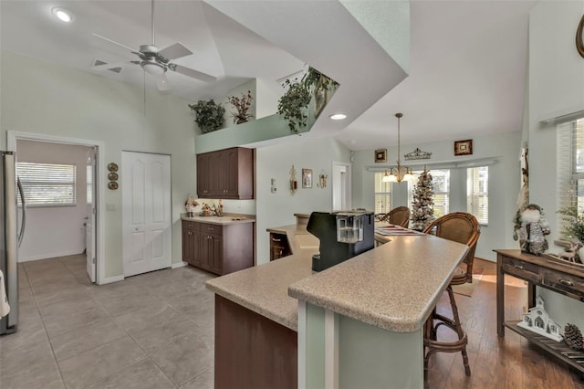 kitchen with high vaulted ceiling, a kitchen breakfast bar, ceiling fan with notable chandelier, stainless steel fridge, and dark brown cabinets