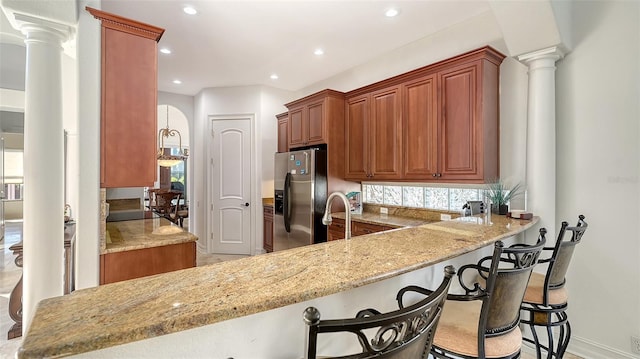 kitchen featuring kitchen peninsula, decorative columns, light stone counters, stainless steel fridge with ice dispenser, and a breakfast bar area