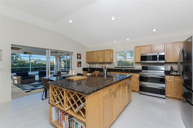 kitchen with lofted ceiling, dark stone counters, light tile patterned floors, an island with sink, and appliances with stainless steel finishes