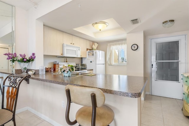 kitchen featuring a raised ceiling, kitchen peninsula, white appliances, cream cabinetry, and light tile patterned floors