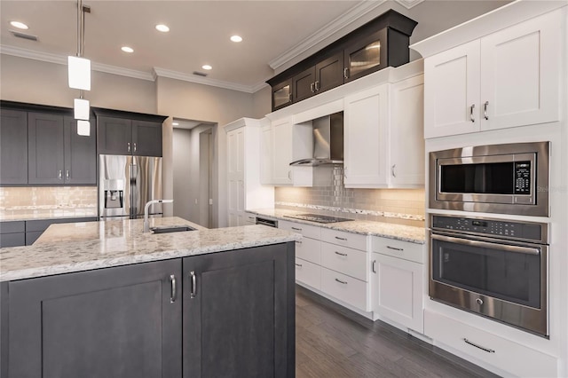 kitchen featuring appliances with stainless steel finishes, wall chimney exhaust hood, a kitchen island with sink, sink, and hanging light fixtures