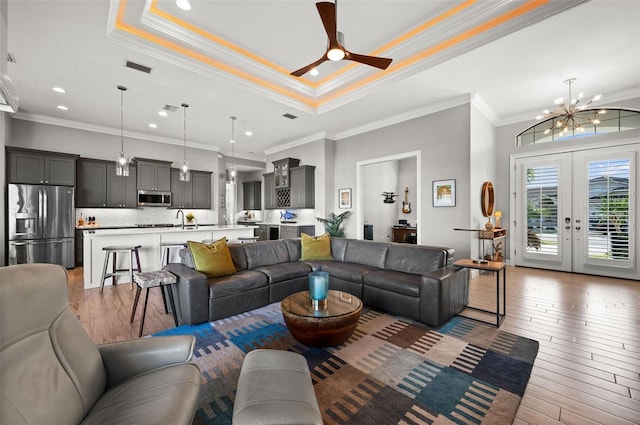 living room featuring french doors, ceiling fan with notable chandelier, light wood-type flooring, ornamental molding, and a tray ceiling