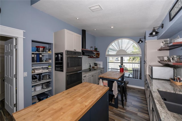 kitchen with stainless steel double oven, wall chimney range hood, dark hardwood / wood-style floors, white cabinetry, and butcher block counters