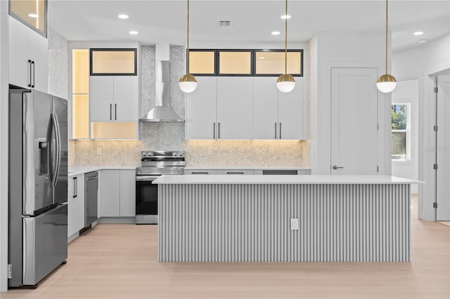 kitchen with appliances with stainless steel finishes, wall chimney range hood, pendant lighting, light hardwood / wood-style floors, and a kitchen island