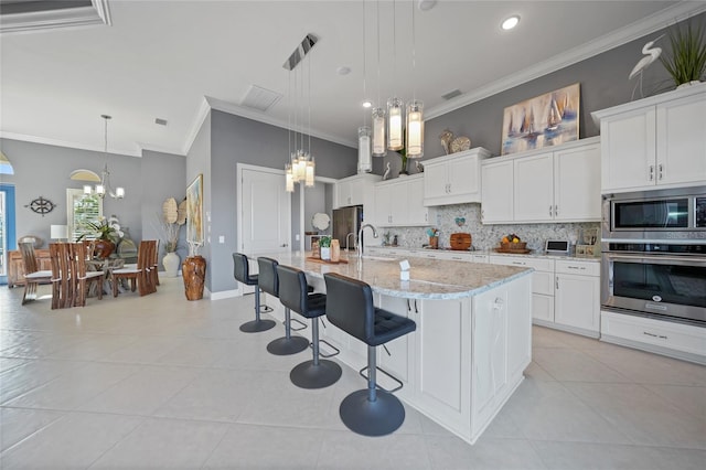 kitchen with white cabinetry, an island with sink, decorative light fixtures, appliances with stainless steel finishes, and ornamental molding