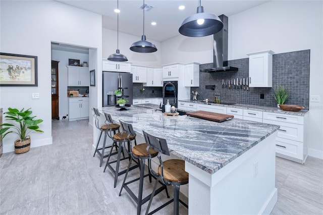 kitchen featuring decorative backsplash, stainless steel fridge, wall chimney exhaust hood, decorative light fixtures, and white cabinetry