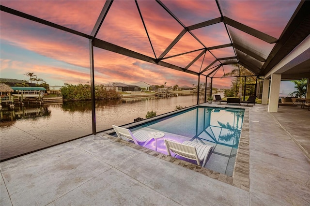 pool at dusk featuring glass enclosure, a water view, and a patio