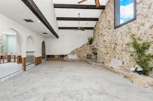 unfurnished living room featuring ceiling fan, a stone fireplace, a high ceiling, and beamed ceiling