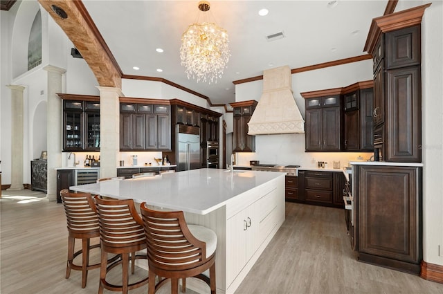 kitchen with dark brown cabinetry, stainless steel appliances, premium range hood, and an island with sink