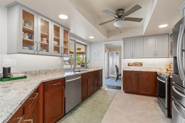 kitchen with sink, ceiling fan, stainless steel appliances, light stone counters, and a tray ceiling