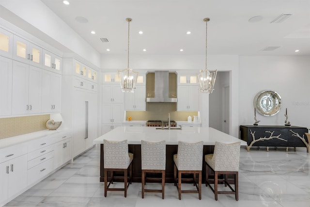 kitchen featuring wall chimney range hood, a kitchen bar, decorative backsplash, a center island with sink, and white cabinets