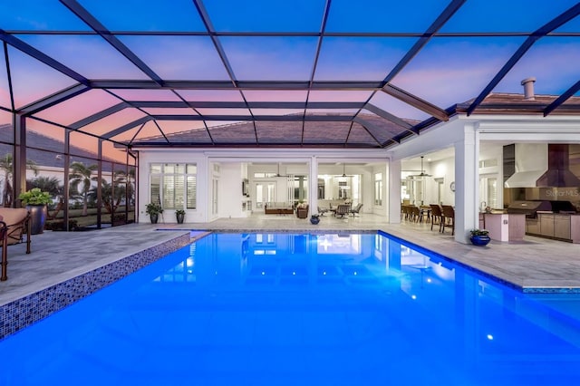 pool at dusk featuring glass enclosure, ceiling fan, a patio area, and exterior kitchen