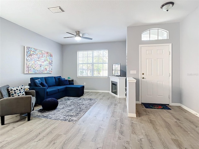 living area with visible vents, light wood-style flooring, a ceiling fan, a glass covered fireplace, and baseboards