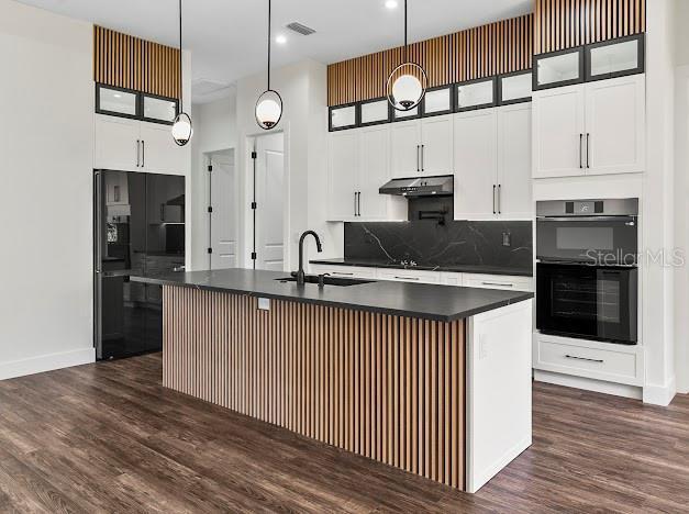 kitchen with black appliances, an island with sink, hanging light fixtures, and dark wood-type flooring