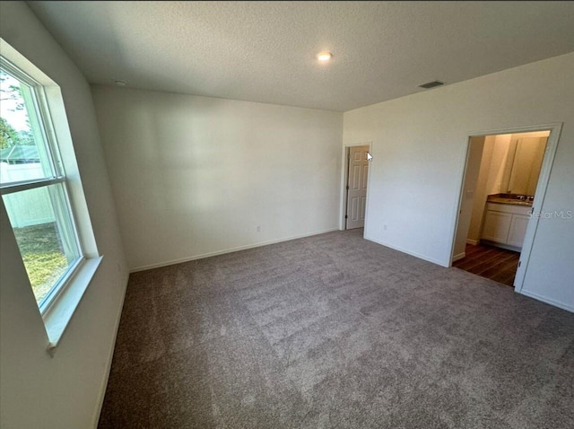 unfurnished bedroom with a textured ceiling, dark colored carpet, visible vents, and ensuite bathroom