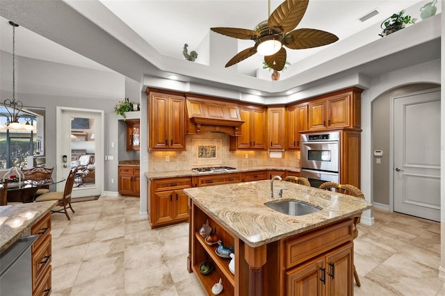 kitchen with light stone countertops, custom exhaust hood, stainless steel appliances, a kitchen island with sink, and sink