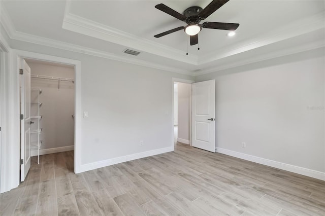 unfurnished bedroom featuring ceiling fan, crown molding, light hardwood / wood-style floors, a tray ceiling, and a walk in closet