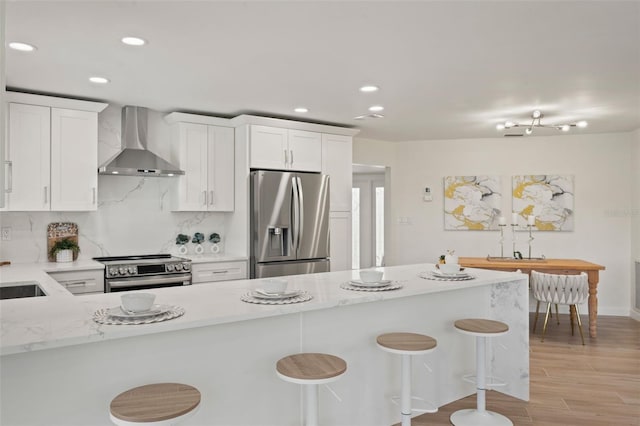 kitchen featuring appliances with stainless steel finishes, white cabinets, tasteful backsplash, a kitchen bar, and wall chimney range hood