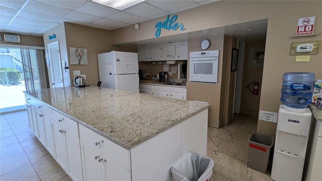 kitchen with a center island, a drop ceiling, light stone counters, white appliances, and white cabinets