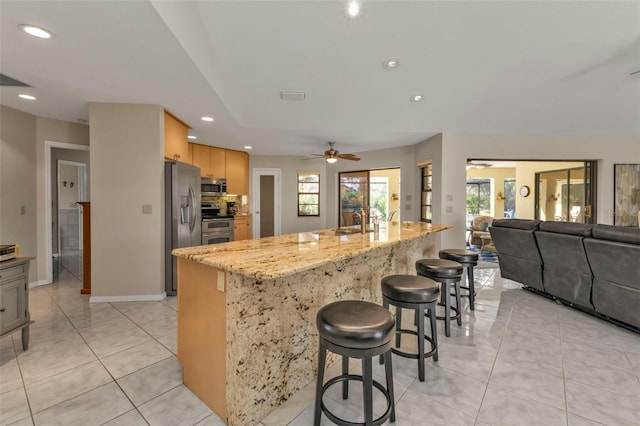 kitchen with appliances with stainless steel finishes, light stone counters, a breakfast bar, ceiling fan, and a kitchen island with sink