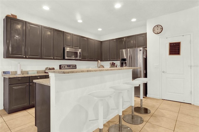 kitchen with appliances with stainless steel finishes, a breakfast bar, dark brown cabinetry, light tile patterned floors, and a center island with sink