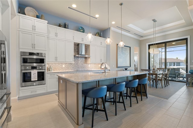 kitchen with white cabinets, stainless steel double oven, a kitchen island with sink, and wall chimney exhaust hood