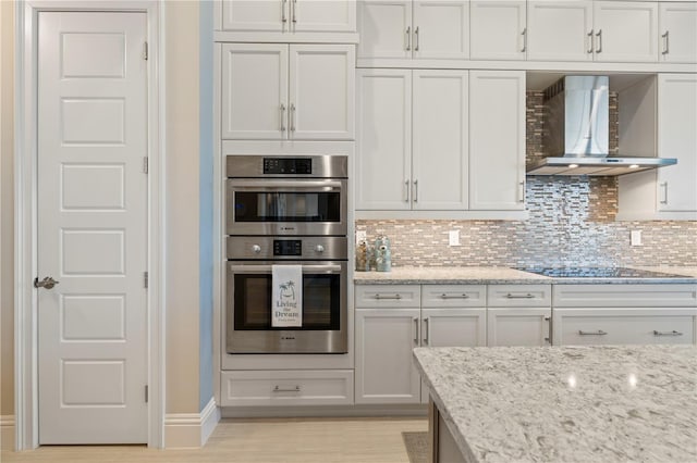 kitchen featuring black electric stovetop, wall chimney range hood, light stone countertops, tasteful backsplash, and stainless steel double oven