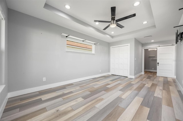 unfurnished bedroom featuring a raised ceiling, ceiling fan, a barn door, light hardwood / wood-style floors, and a closet