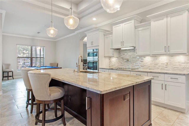 kitchen featuring a center island with sink, hanging light fixtures, double oven, tasteful backsplash, and white cabinetry