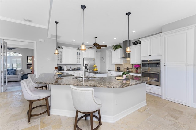 kitchen with wall chimney exhaust hood, decorative light fixtures, a large island with sink, dark stone countertops, and sink