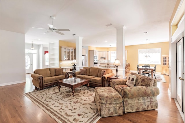 living room featuring built in shelves, wood-type flooring, ceiling fan with notable chandelier, and decorative columns