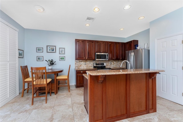 kitchen with appliances with stainless steel finishes, backsplash, light stone counters, and a kitchen island with sink