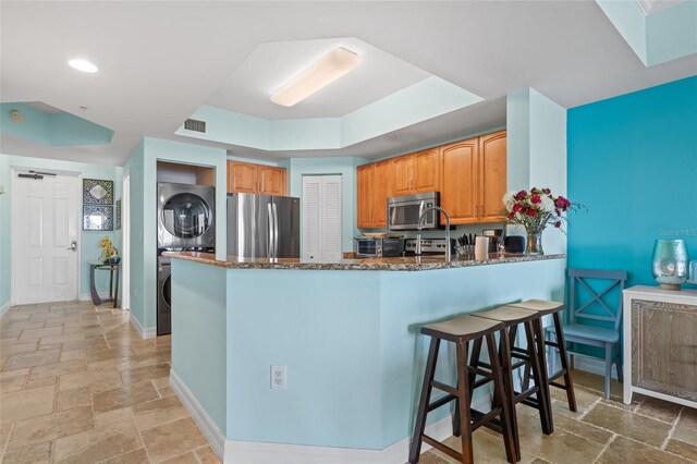 kitchen featuring dark stone counters, stacked washer and clothes dryer, a kitchen breakfast bar, kitchen peninsula, and stainless steel appliances