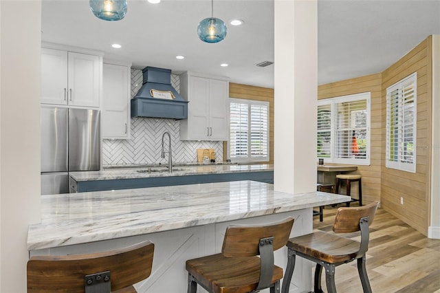 kitchen with pendant lighting, custom exhaust hood, stainless steel fridge, light stone countertops, and white cabinetry