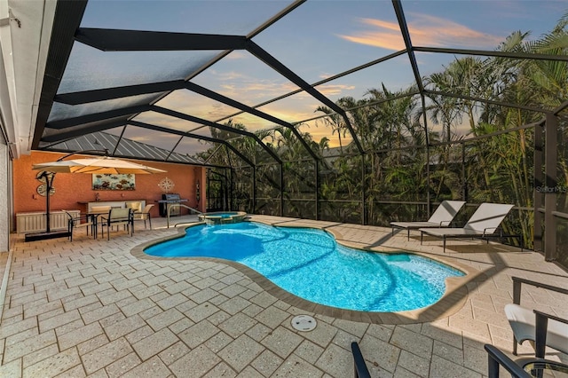 pool at dusk with an in ground hot tub, area for grilling, glass enclosure, and a patio area