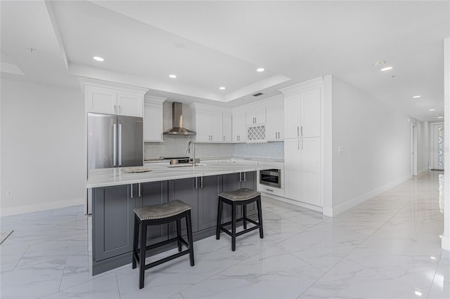 kitchen with wall chimney range hood, stainless steel refrigerator, a kitchen island with sink, a kitchen breakfast bar, and white cabinets