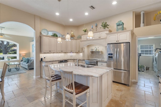 kitchen with sink, hanging light fixtures, stainless steel appliances, light stone countertops, and a center island with sink