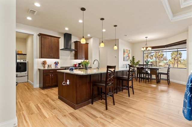 kitchen with wall chimney exhaust hood, light stone counters, hanging light fixtures, light wood-type flooring, and a kitchen island with sink