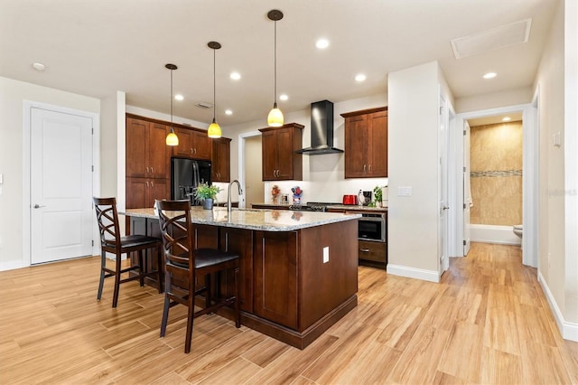 kitchen featuring decorative light fixtures, black refrigerator with ice dispenser, an island with sink, light stone countertops, and wall chimney range hood