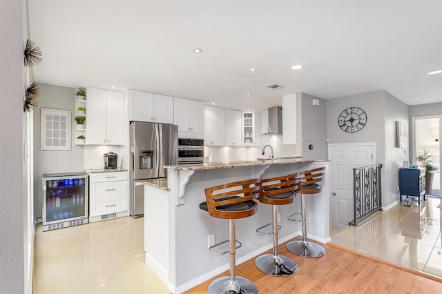 kitchen with white cabinetry, beverage cooler, wall chimney range hood, kitchen peninsula, and appliances with stainless steel finishes