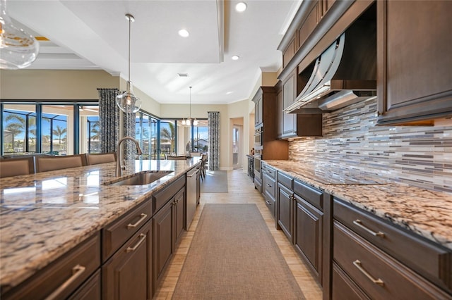 kitchen featuring wall chimney range hood, sink, hanging light fixtures, light stone countertops, and dark brown cabinets