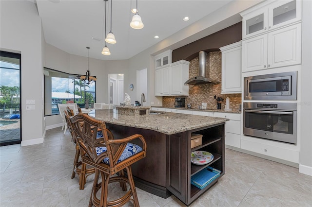 kitchen with wall chimney range hood, hanging light fixtures, a kitchen island with sink, appliances with stainless steel finishes, and white cabinets