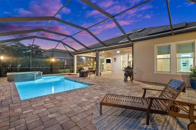 pool at dusk with an in ground hot tub, glass enclosure, an outdoor fire pit, and a patio area