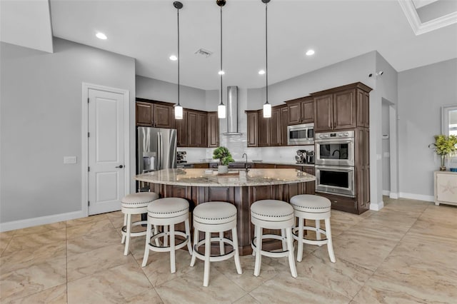 kitchen with decorative light fixtures, an island with sink, stainless steel appliances, light stone countertops, and wall chimney range hood