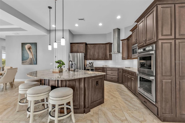 kitchen with appliances with stainless steel finishes, a kitchen island with sink, a kitchen breakfast bar, light stone counters, and wall chimney exhaust hood