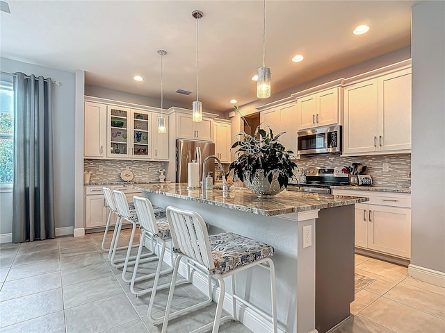 kitchen with light stone counters, stainless steel appliances, a kitchen island with sink, pendant lighting, and light tile patterned flooring