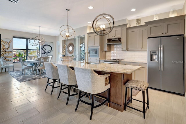 kitchen with a breakfast bar, appliances with stainless steel finishes, gray cabinets, light stone countertops, and a kitchen island with sink