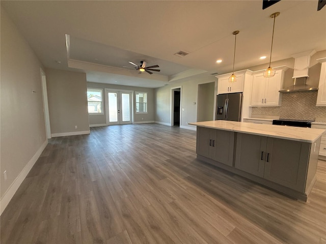 kitchen featuring stainless steel refrigerator with ice dispenser, a center island, a raised ceiling, white cabinets, and wall chimney range hood